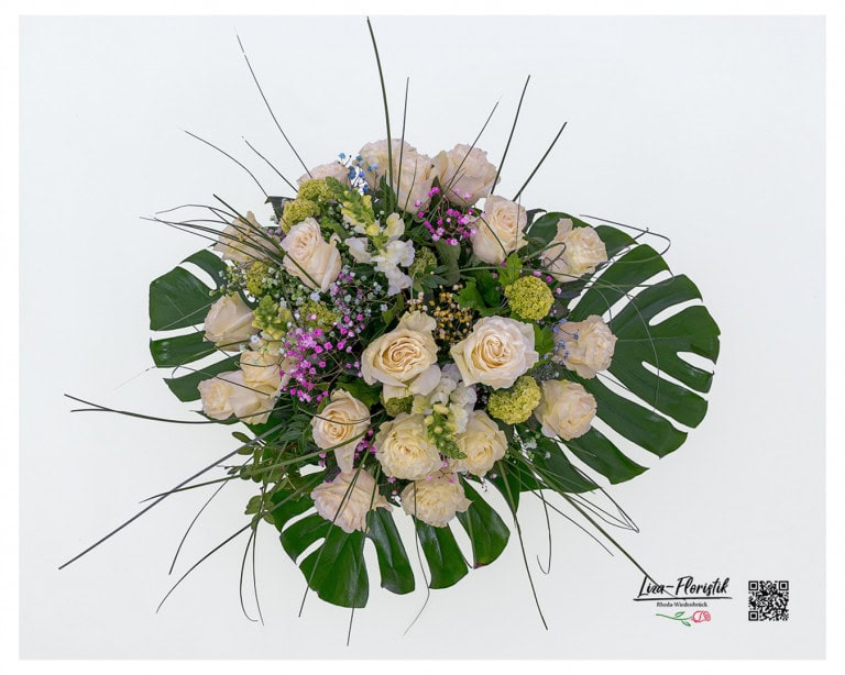 Large bouquet of flowers in top view, artfully tied with 20 creamy white Ecuador roses of the Mondial variety, seasonal viburnum, white snapdragons and colored gypsophila in pink, purple and blue. Framed by decorative leafy green, including large monstera leaves and delicate grasses for a tropical, elegant look.